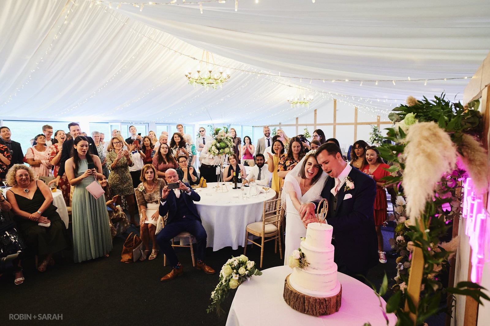 Bride and groom cut wedding cake in marquee at Wethele Manor as guests watch and take photos