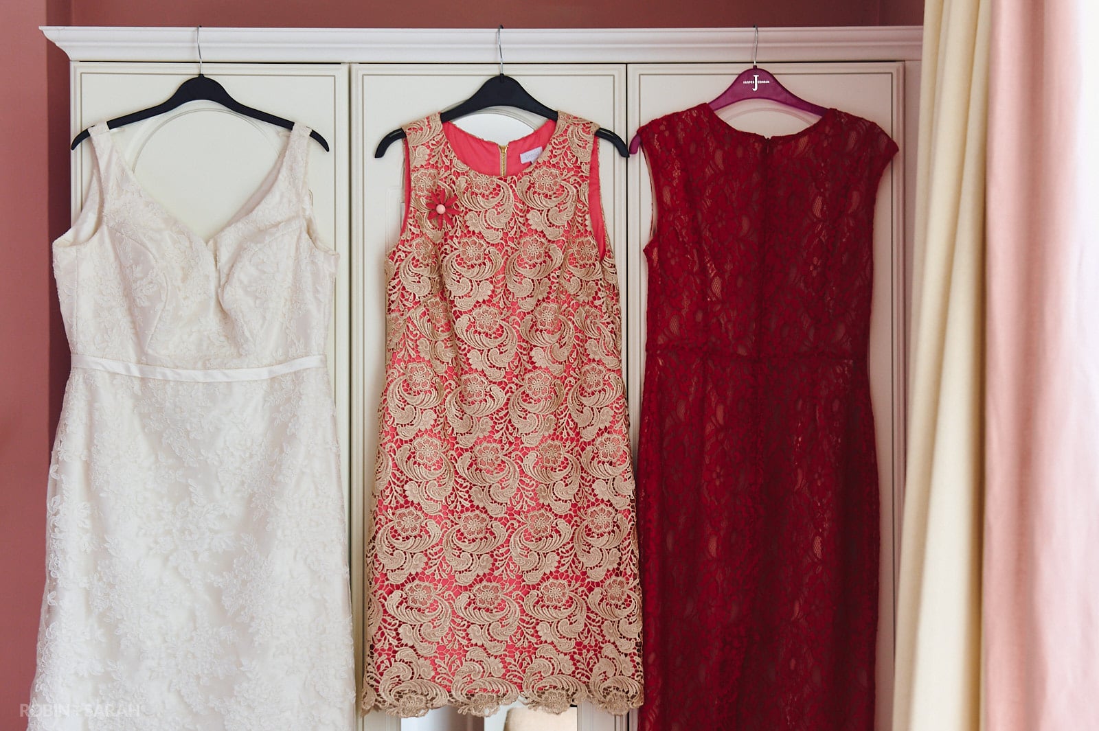 Short white wedding dress and two red bridesmaids dresses hang up on wardrobe