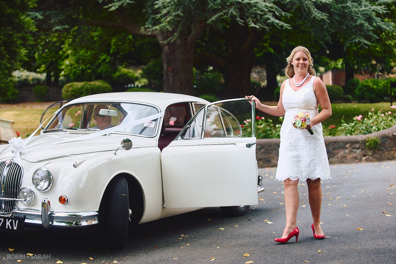 Bride in knee-length white wedding dress arrives in old Jaguar car