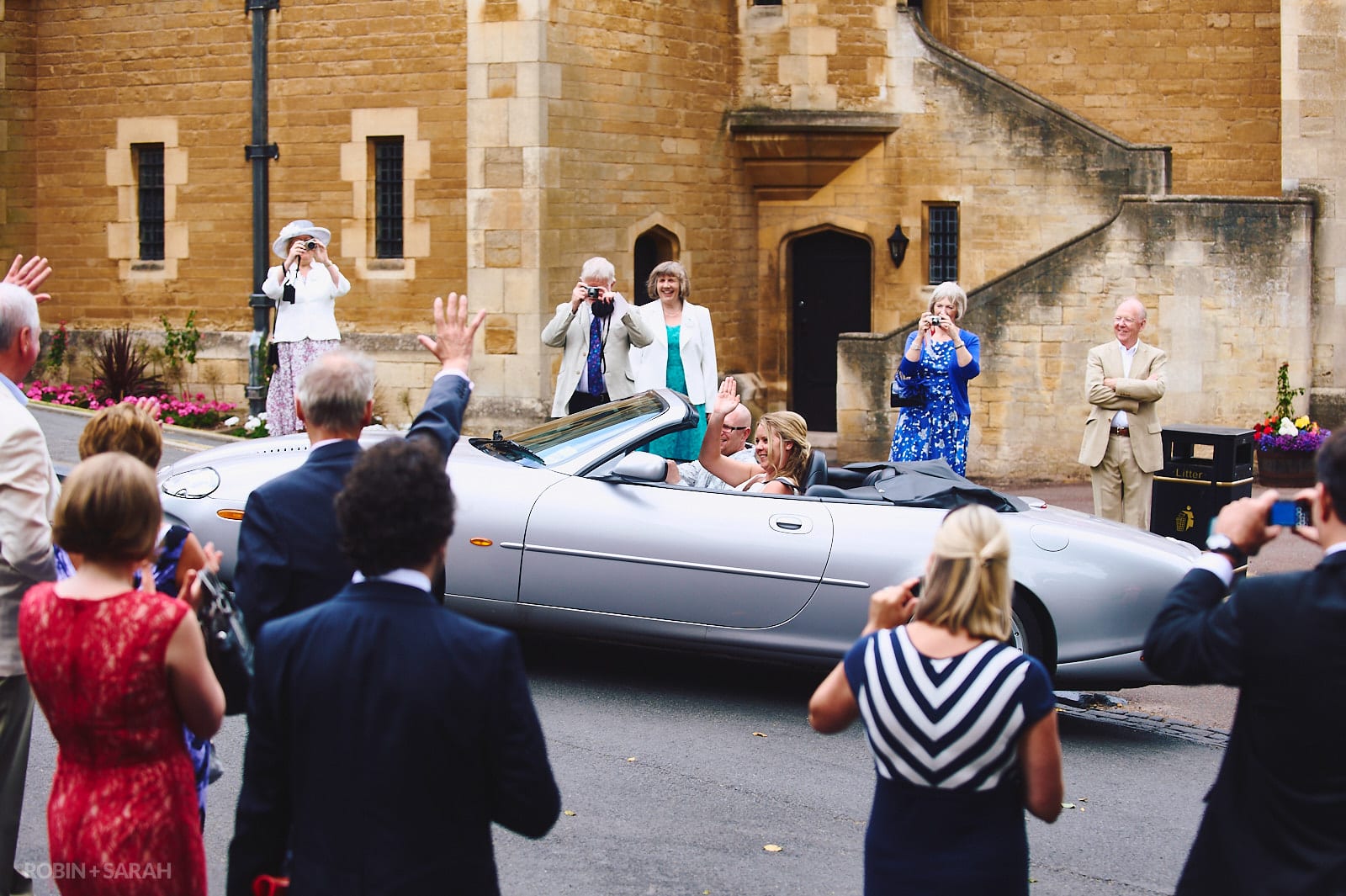 Bride and groom drive off in open-top car as guests wave and take pictures