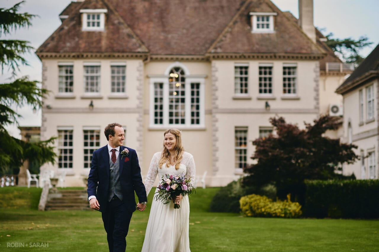 Bride and groom walk through beautiful gardens with Brockencote Hall in background