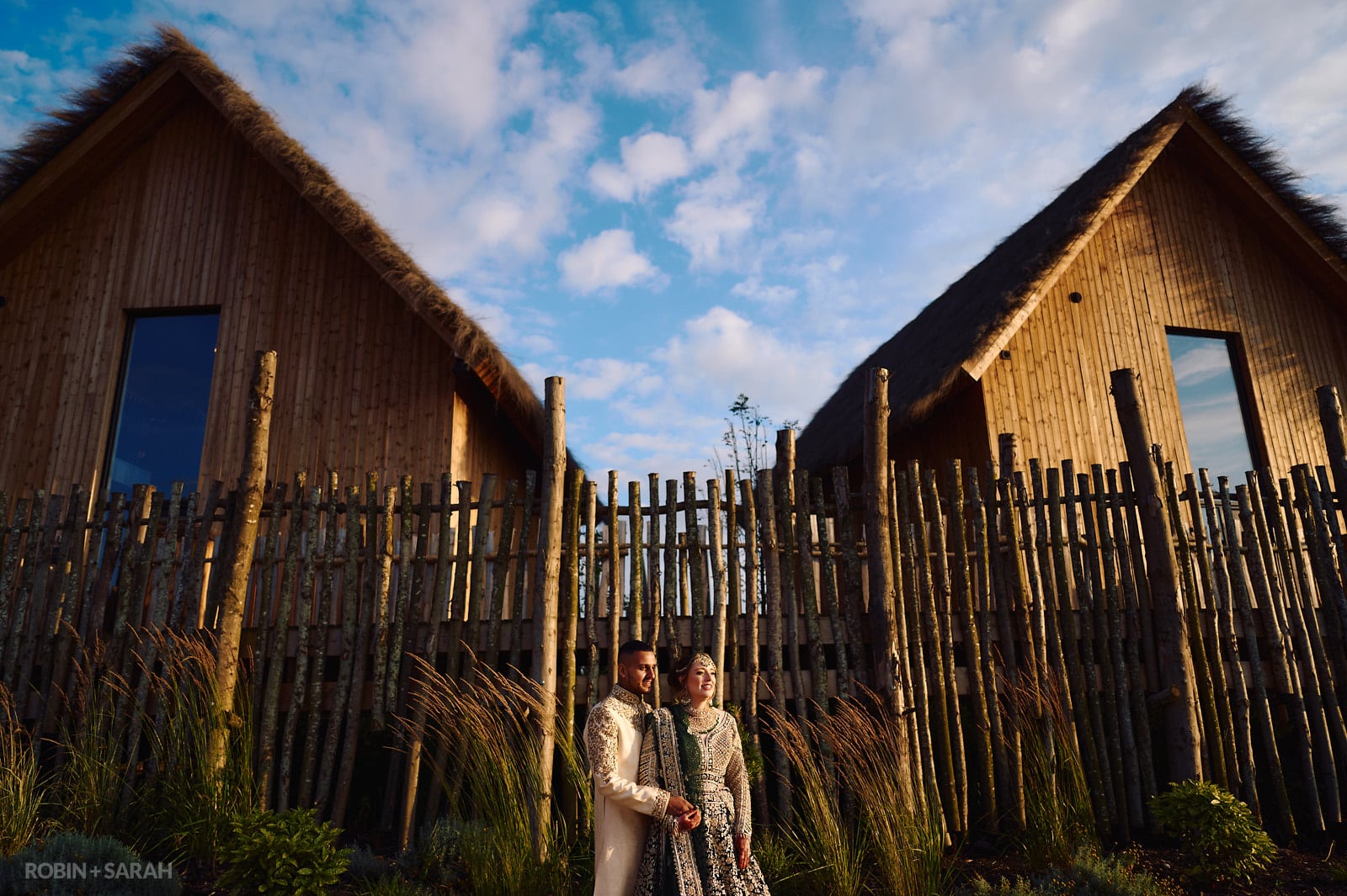 Bride and groom in Indian wedding attire stand between two wooden lodges at West Midlands Safari Park