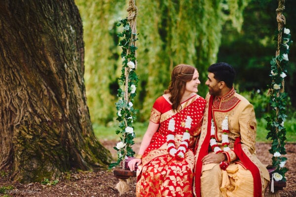 Bride and groom in Indian wedding outfits sit on swing under old tree at Stanbrook Abbey