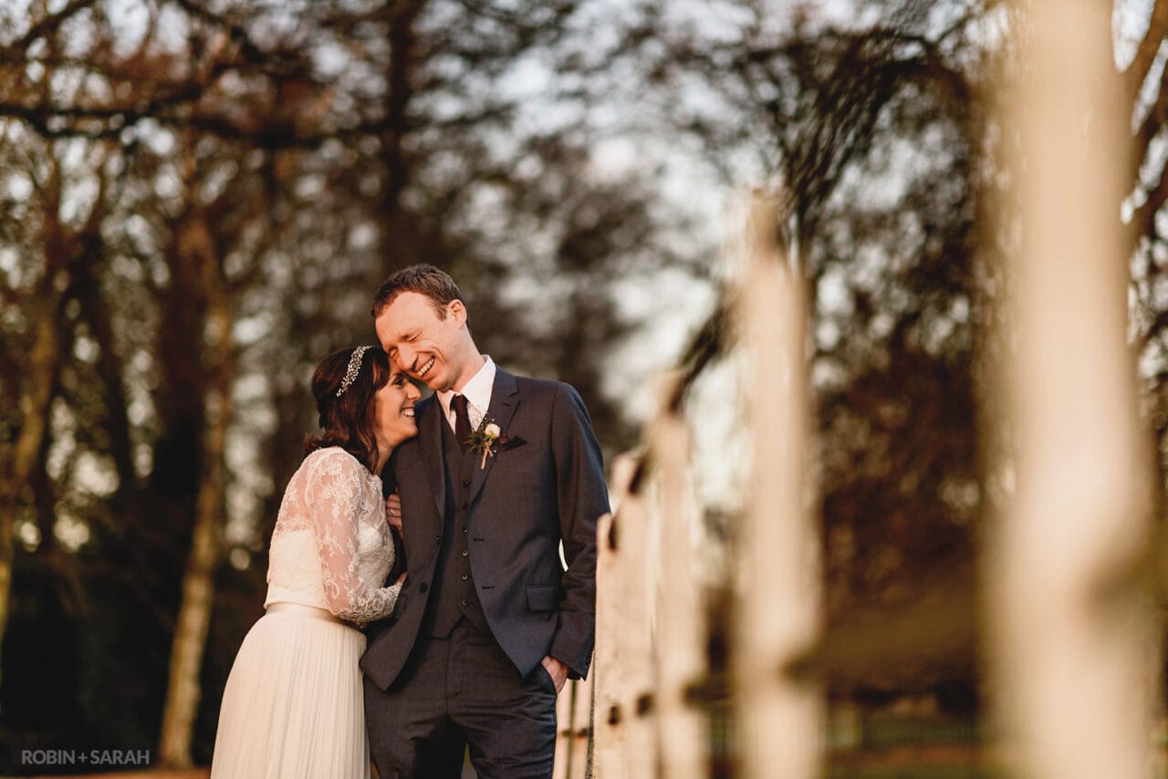 Bride and groom laugh together as they lean on a fence in soft evening light