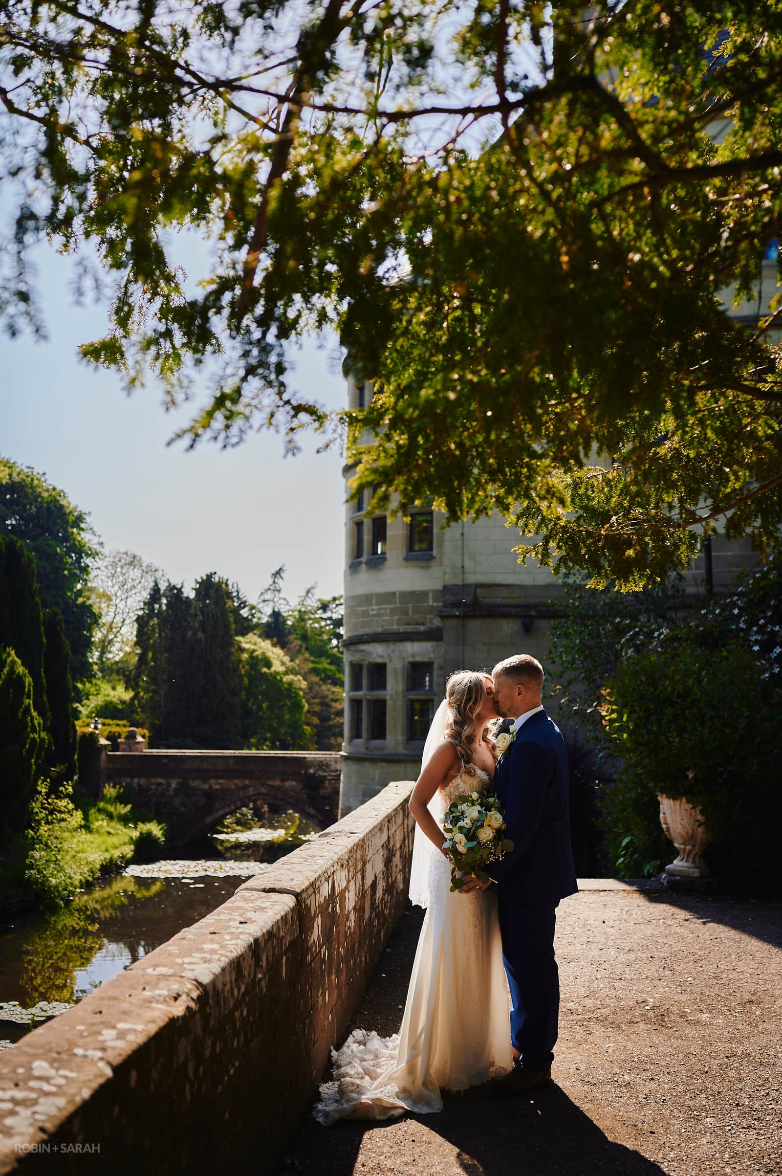 Bride and groom kiss under tree on terrace at Coombe Abbey