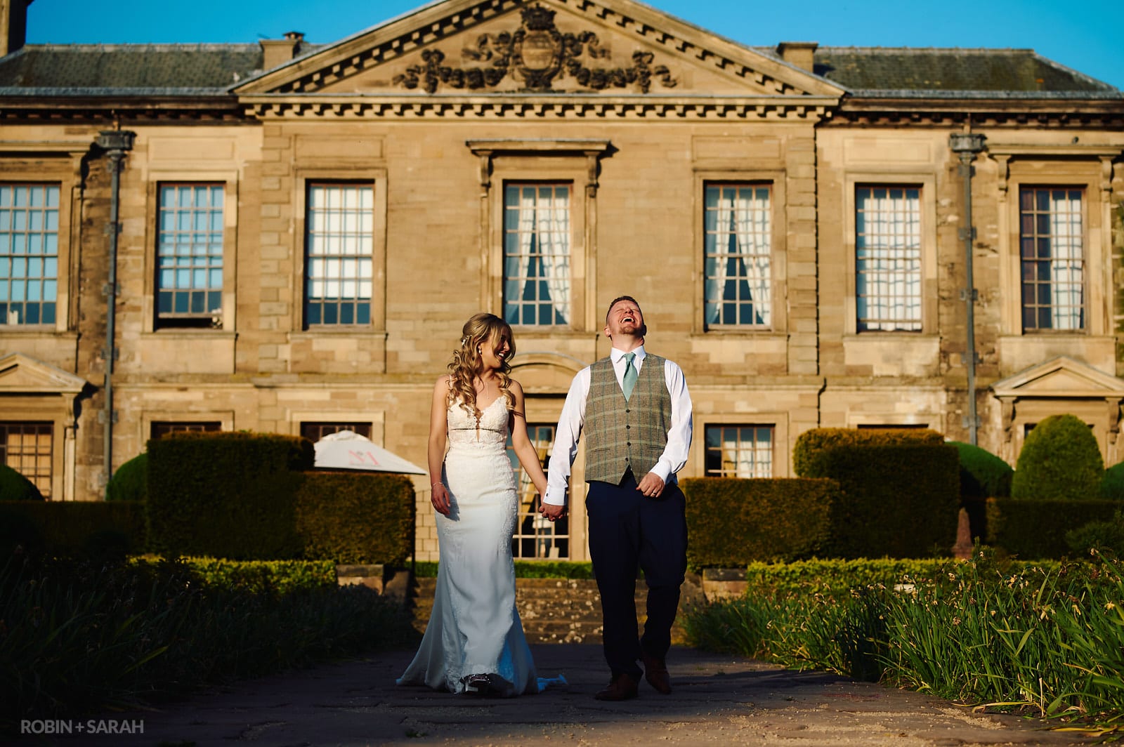 Bride and groom laughing as they walk along path at Coombe Abbey