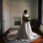 Portrait of beautiful bride in elegant wedding dress standing in a spartan room in window light