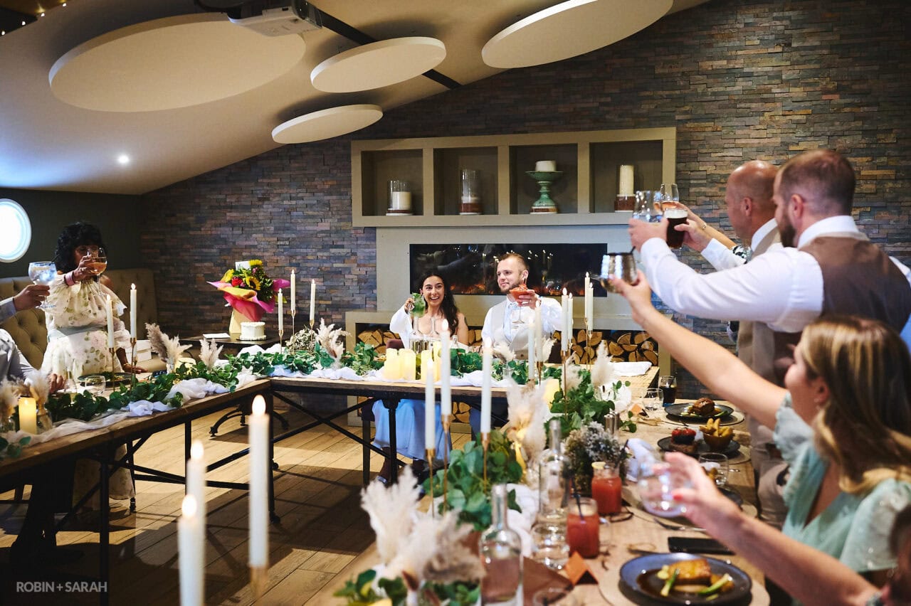 Bride and groom sit down for dinner at The Barn at Berryfields as wedding guests raise a toast