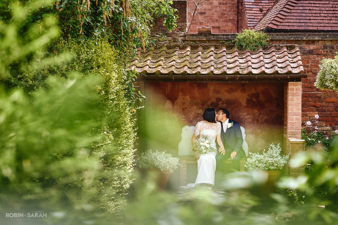 Bride and groom kiss while sitting in alcove in beautiful gardens