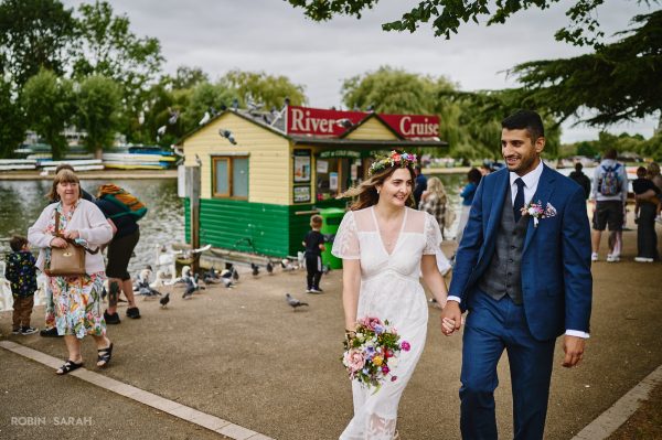 Bride and groom walk hand in hand alongside a river as members of the public watch