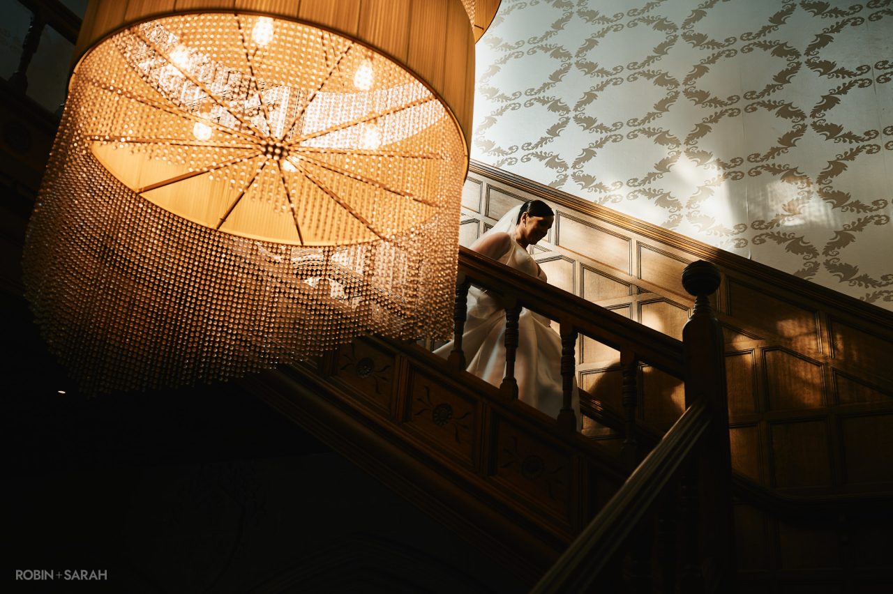 Bride walks down the staircase at Hampton Manor, with the large chandelier and moody lighting