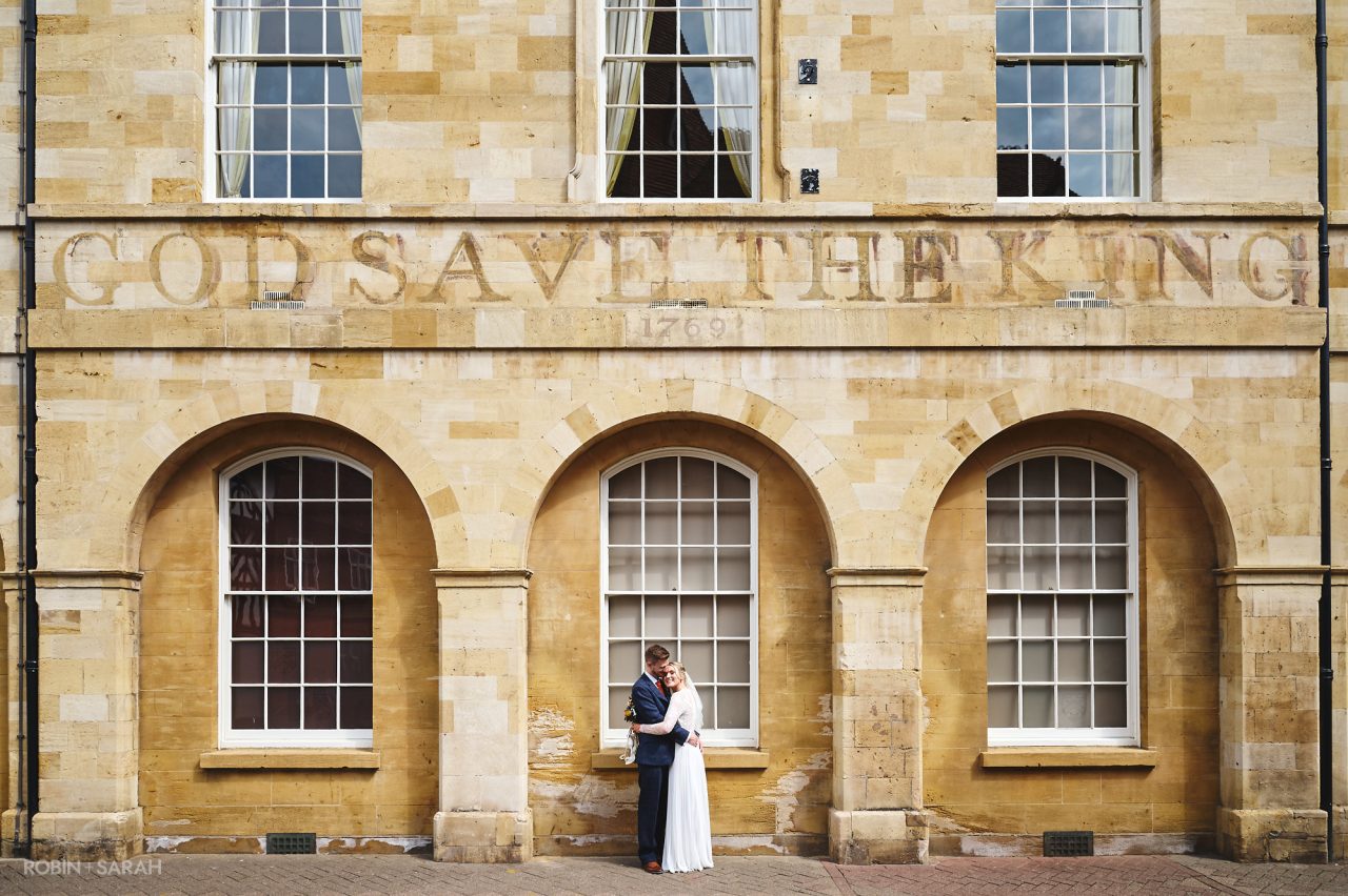 Newly married couple hug under one of the arches of the Town Hall in Stratford-upon-Avon, with the words God Save The King faded on the wall above