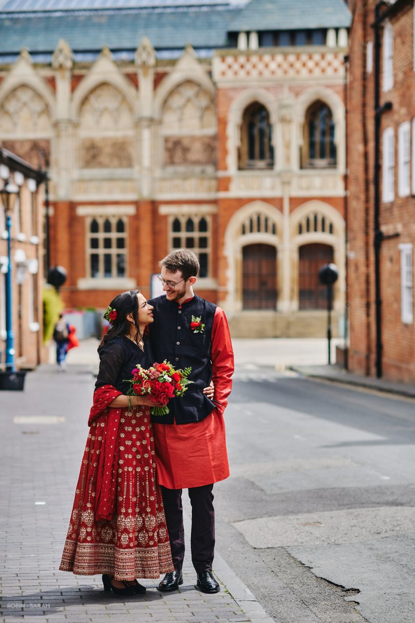 Newly married couple wearing red and black Indian wedding attire stand in front of theatre in Stratford-upon-Avon