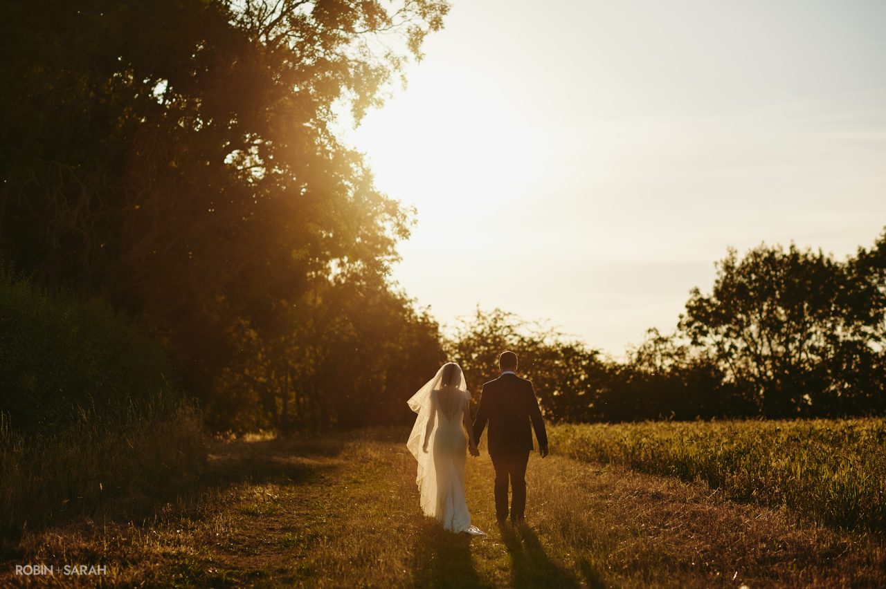 Bride and groom walk through fields at sunset, at Wethele Manor near Leamington Spa, Warwickshire