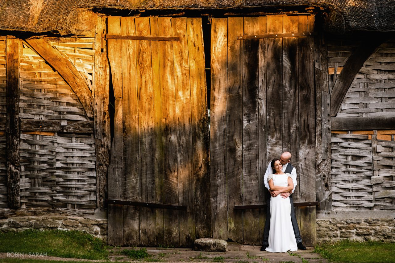 Bride and groom cuddle while leaning against a beautiful old barn at Avoncroft Museum in Bromsgrove