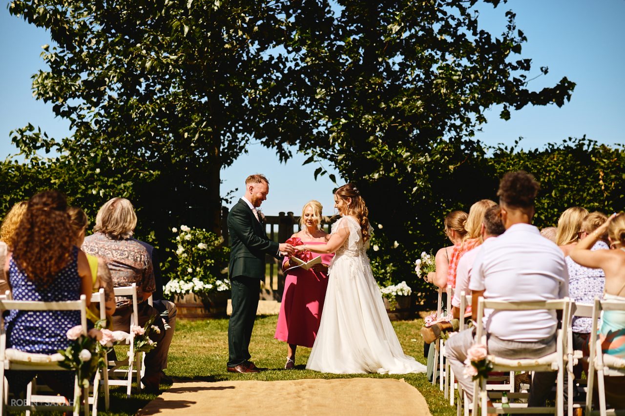 Bride and groom exchange wedding rings during outdoor ceremony in the gardens of Charingworth House on a sunny day